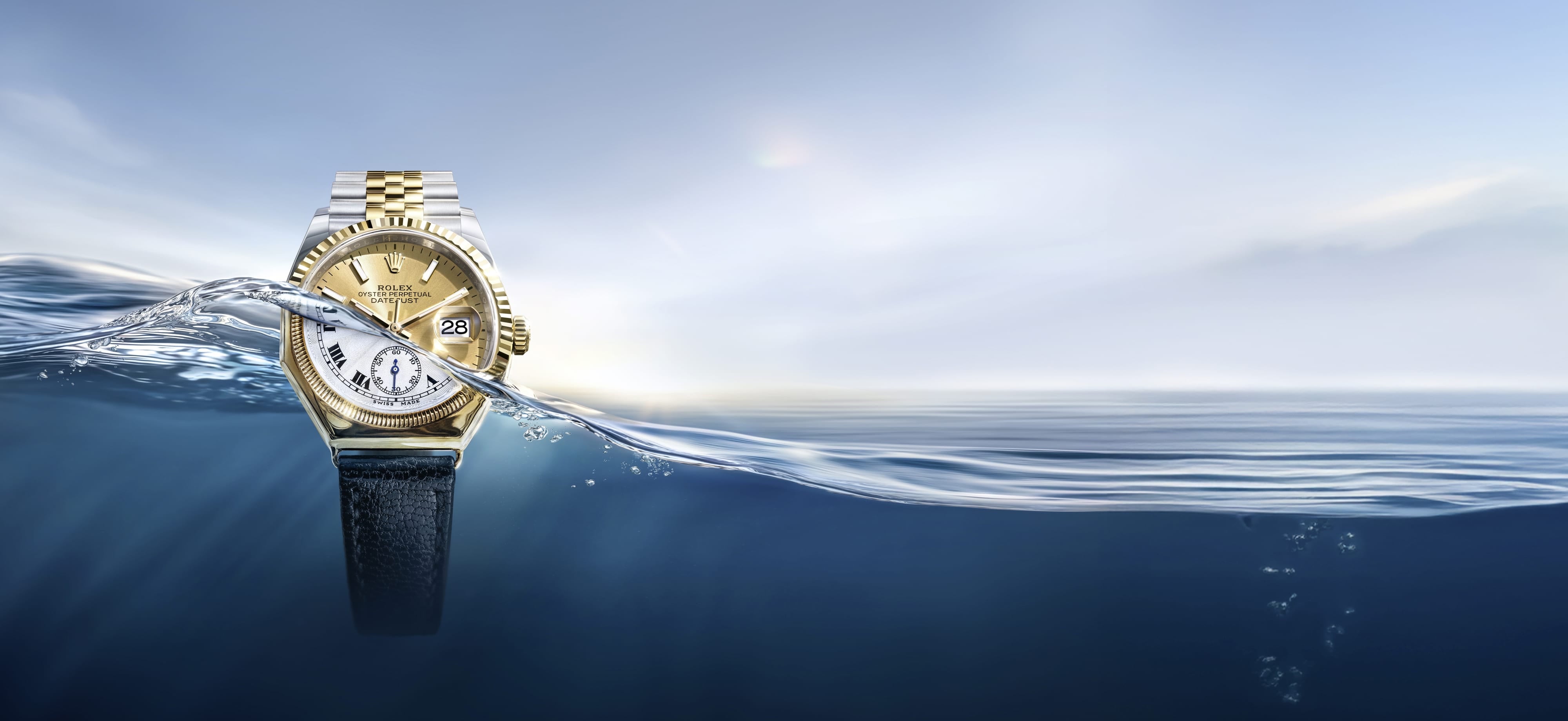 A vintage gold Rolex Oyster watch with a black strap partially submerged in water. In the clear sky above the wave emerges a modern Rolex Oyster Perpetual Datejust in yellow Rolesor with a bracelet made of Oystersteel and solid 18 carat yellow gold.
