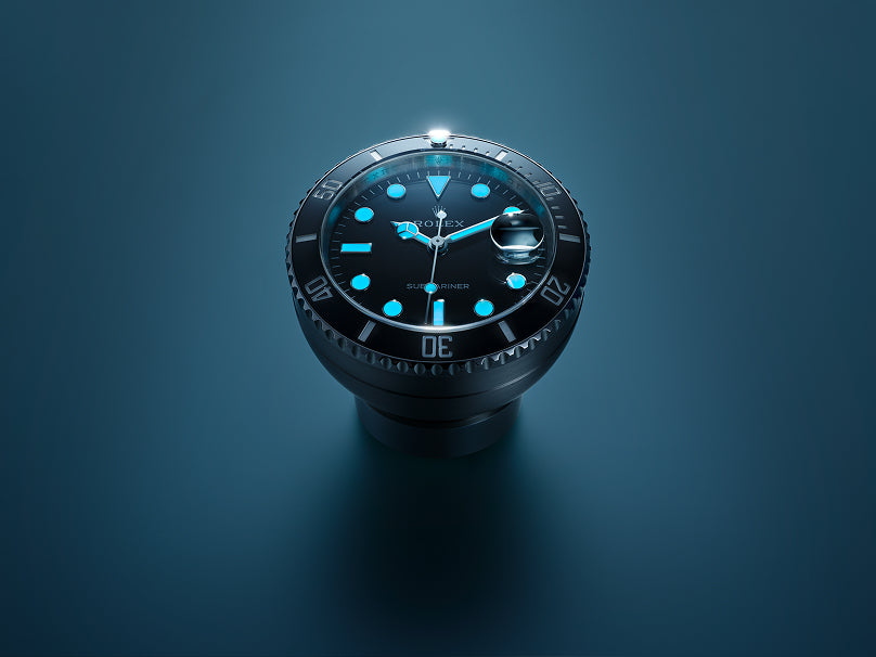 A Rolex Submariner desk clock in Oystersteel, with black dial and black Cerachrom ceramic bezel, on a muted blue surface with soft lighting. The photo-luminescent hands and indices are emitting a bluish glow.
