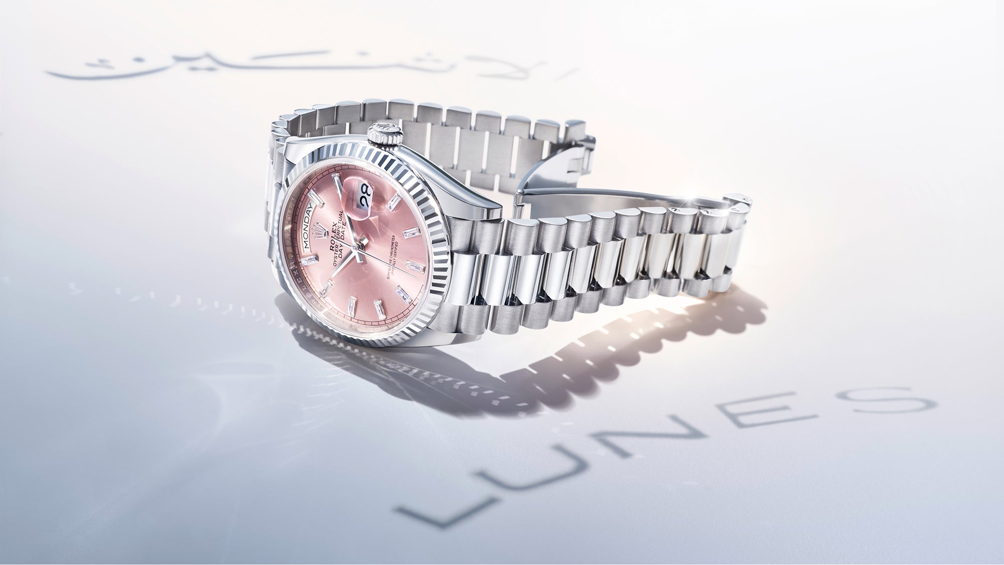 A Rolex Day-Date in solid 18 carat white gold with a pink dial, set on a light grey background, with days of the week in different languages reflected on the surface.