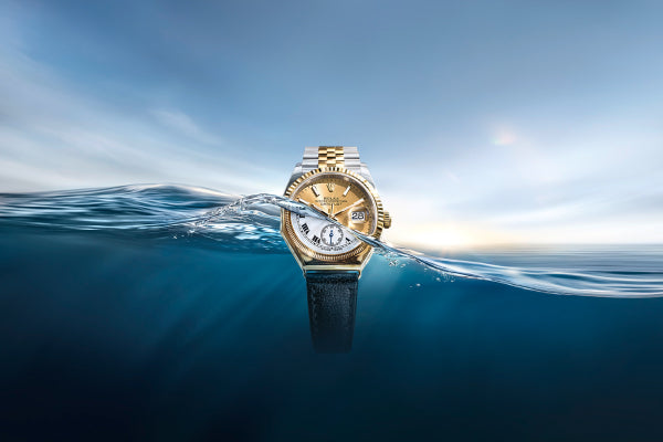 A vintage gold Rolex Oyster watch with a black strap partially submerged in water. In the clear sky above the wave, emerges a modern Rolex Oyster Perpetual Datejust in yellow Rolesor with a bracelet made of Oystersteel and solid 18 carat yellow gold.