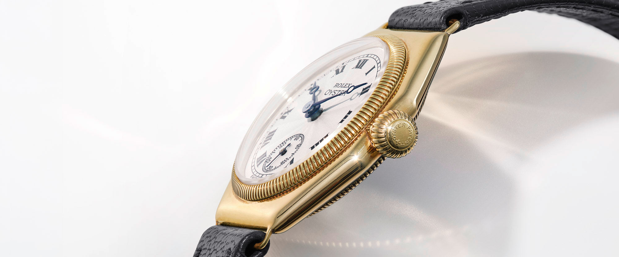 A vintage, gold Rolex Oyster wristwatch with a white face, fitted to a black leather strap, set against a light gray background.