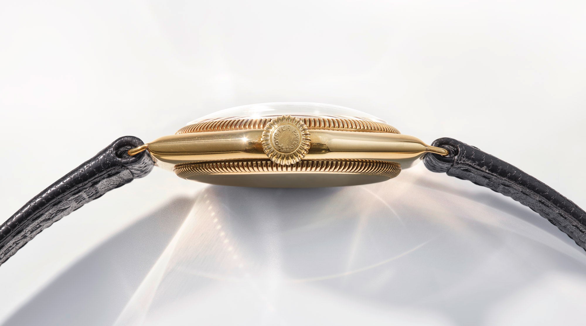 A profile view of a vintage, gold Rolex Oyster watch, prominently featuring the screw-down crown, which has the words "Oyster Patented" embossed on it in a circle.