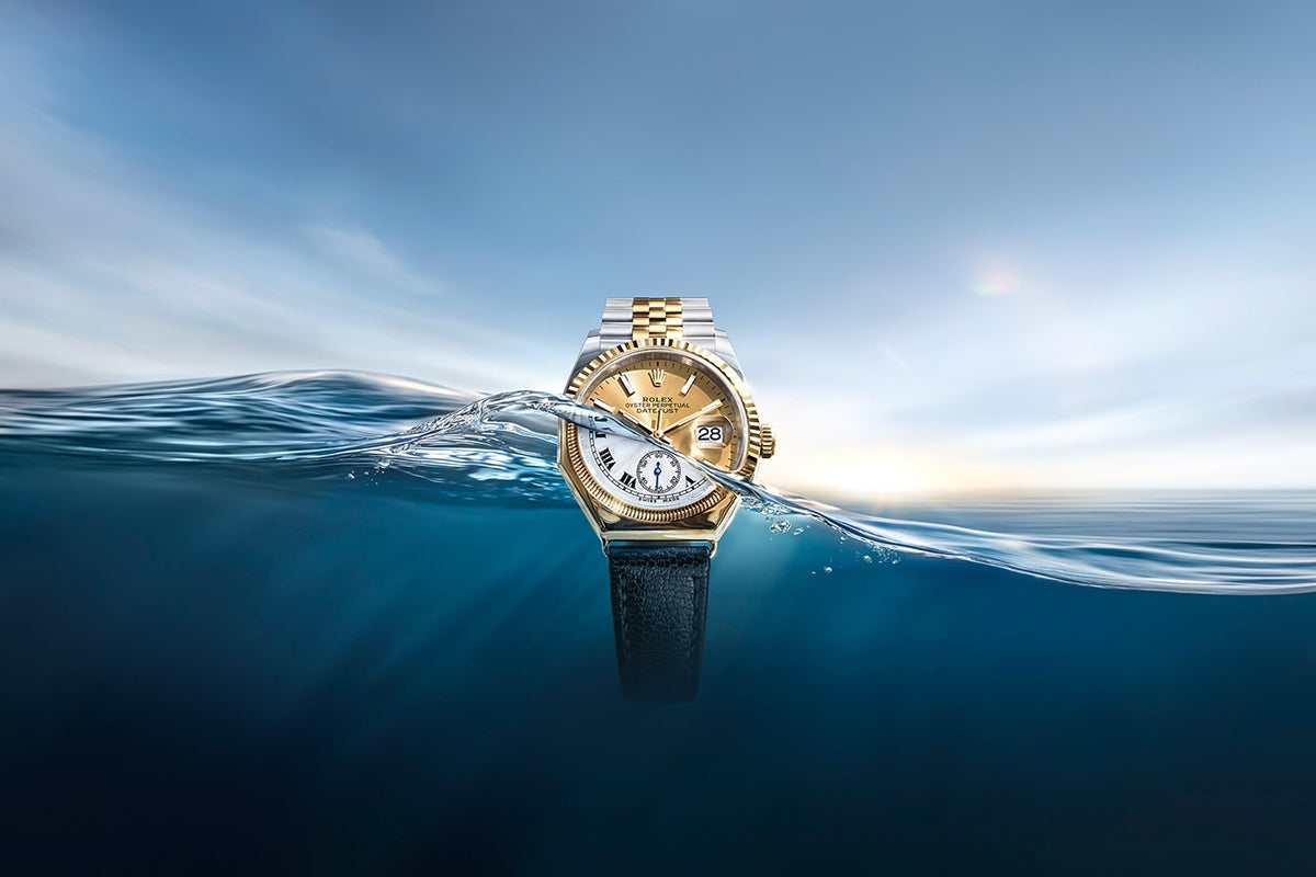 A vintage gold Rolex Oyster watch with a black strap partially submerged in water. In the clear sky above the wave emerges a modern Rolex Oyster Perpetual Datejust in yellow Rolesor with a bracelet made of Oystersteel and solid 18 carat yellow gold.
