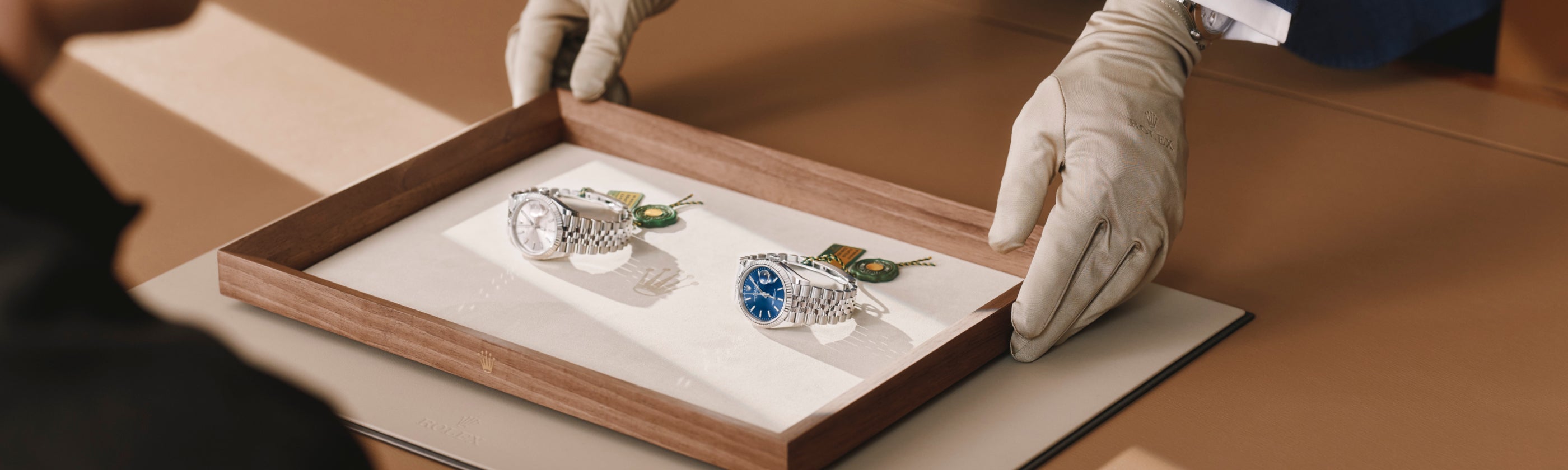 A Luxury Watch Specialist, wearing gloves, presenting a finely-crafted tray with two Rolex Datejust timepieces on it to a client sitting at a wooden table.