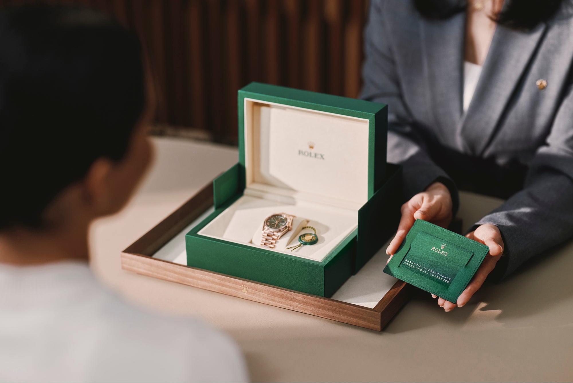 A Luxury Watch Specialist presenting a Rolex International Guarantee to a client, alongside a new Rolex in its box on a finely-crafted tray, to a client sitting across from them at a wooden table.