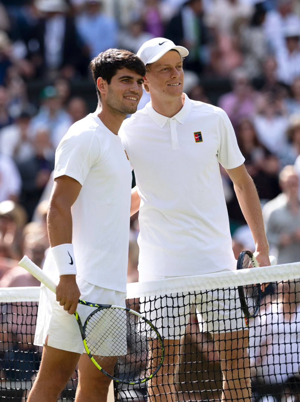 Two tennis players in white uniforms standing behind a net with a blurred crowd in the background.