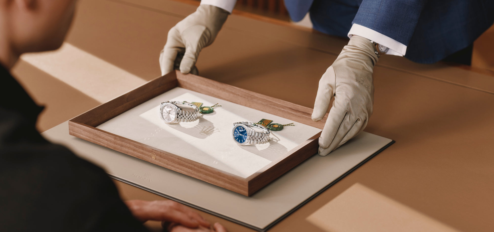A Luxury Watch Specialist, wearing gloves, presenting a finely-crafted tray with two Rolex Datejust timepieces on it to a client sitting at a wooden table.